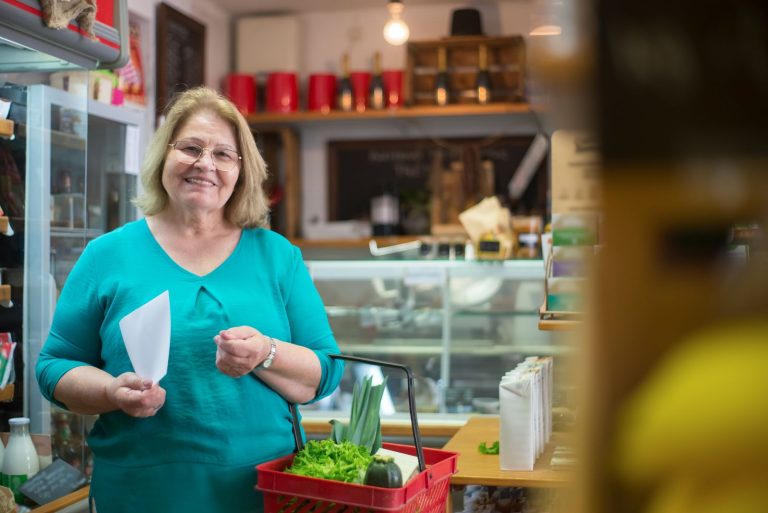 Une femme souriante tient une liste de courses et un panier rempli de légumes dans une épicerie
