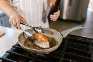 Une personne retourne un filet de poisson avec une pince dans une poêle sur une plaque de cuisson