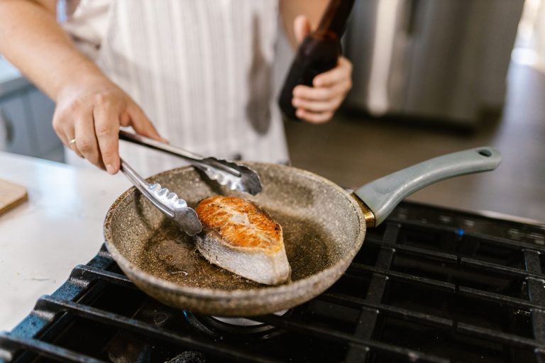 Une personne retourne un filet de poisson avec une pince dans une poêle sur une plaque de cuisson