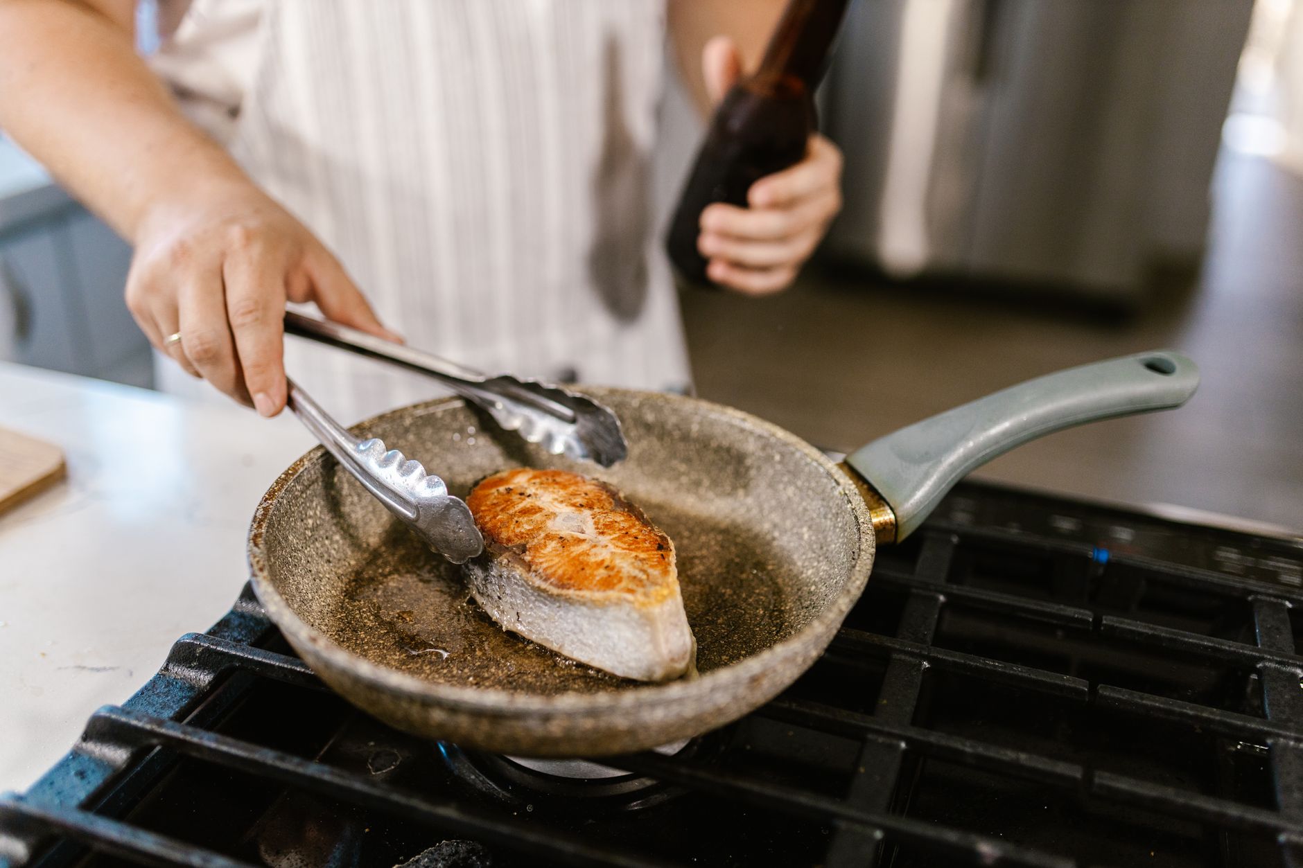 Une personne retourne un filet de poisson avec une pince dans une poêle sur une plaque de cuisson