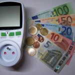 Un wattmètre branché sur une prise française posé à côté de billets et de pièces en euros