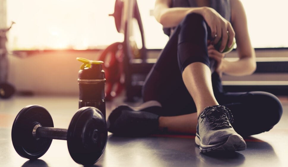 Femme assise au sol en salle de musculation, visage non visible, près d'haltères